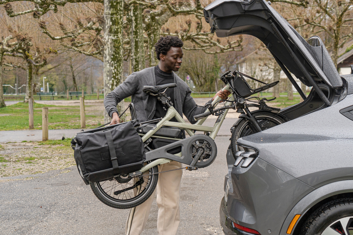 A man carries a folded Tern HSD electric bicycle with a large black pannier bag next to an open trunk of a gray SUV in a park setting.